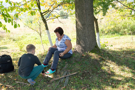 In a serene outdoor setting, a woman and a child enjoy a quiet moment under the shade of a tree. The scene is bathed in natural light, with green grass covering the ground.の写真素材
