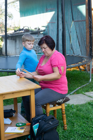 A young boy stands alongside his grandmother as she sits on a small stool, both peering intently at a document on a table outdoors, with a trampoline visible in the backdrop.の写真素材
