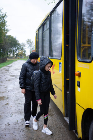 A grandmother and granddaughter are pictured next to a yellow bus. They are standing on a muddy road, and the weather appears to be overcast. The girl is about to board the bus.の写真素材