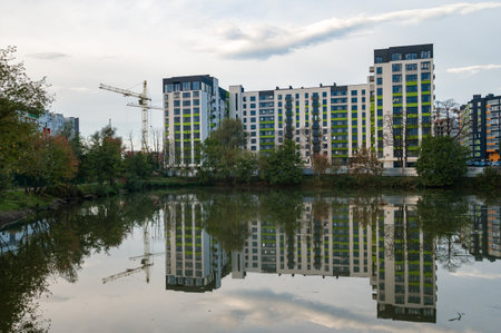 A serene urban pond captures the reflections of contemporary buildings, while nearby trees sway gently in the breeze during a cloudy afternoon.の写真素材