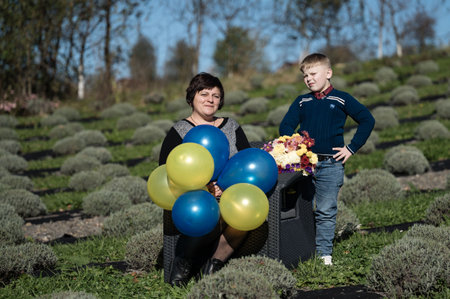 A woman sits on a chair in a lavender field, holding vibrant balloons in blue and yellow while a young boy stands beside her with a bouquet of flowers, radiating happiness.の写真素材