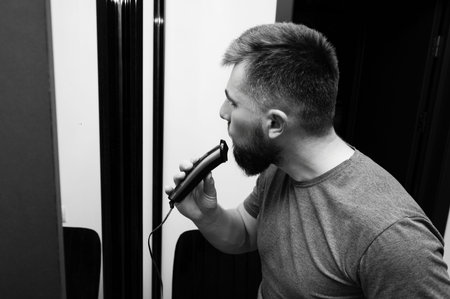 A man focuses intently while trimming his beard with an electric trimmer, immersed in his grooming routine within a stylish interior, showcasing self-care.の写真素材
