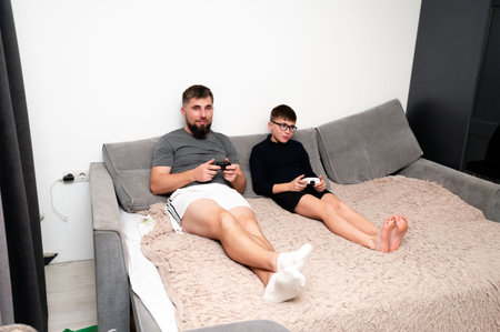 A father and son sit side by side on a large couch, engrossed in gaming. The cozy room features neutral tones, soft blankets, and playful energy as they share this quality time.の写真素材