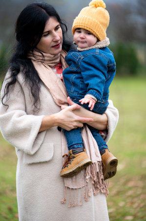 A loving mother, wearing a scarf and coat, tenderly holds her baby outside. The child wears a hat and denim jacket. Both share a moment of warmth and connection.の写真素材