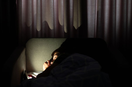 A cozy atmosphere envelops a room as a young person lies on a couch, captivated by the glow of a mobile device amidst gentle shadows and dim light.の写真素材