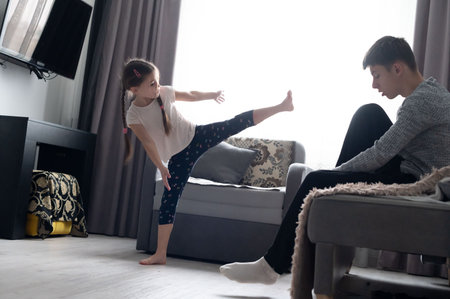 A young girl confidently demonstrates her karate skills in a warm living room, while her father attentively watches, highlighting their playful interaction and family connection.の写真素材
