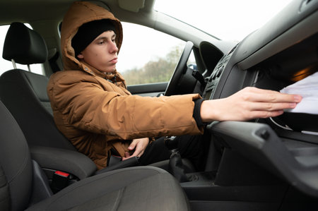 In a cozy vehicle, a young man dressed warmly explores the glove compartment, navigating through papers as he seeks something important during a cold day.の写真素材
