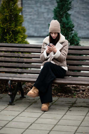 A young woman sits comfortably on a bench wearing a warm coat and hat, absorbed in her phone while enjoying a tranquil afternoon in a peaceful park setting.の写真素材