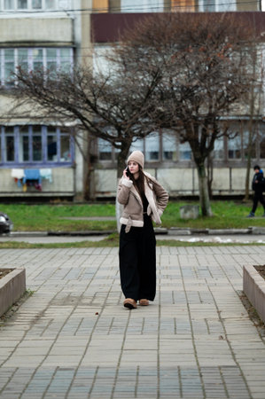 A young woman walks leisurely along a paved path in an urban park, wearing warm winter clothes, while trees without leaves frame her surroundings on a chilly day.の写真素材