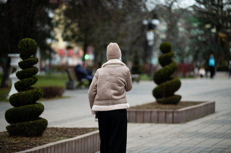 An elderly woman in a warm coat walks leisurely down a tranquil park pathway, flanked by beautifully sculpted bushes under a soft evening sky.の写真素材