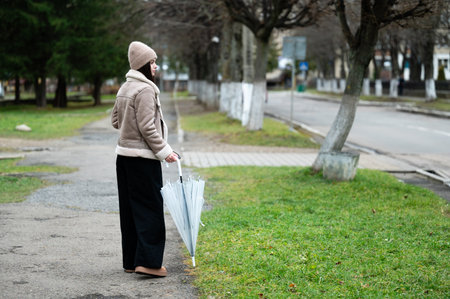 A solitary woman waits on a quiet street, dressed warmly in a soft jacket and hat, holding an umbrella, while the gray clouds suggest impending rain.の写真素材