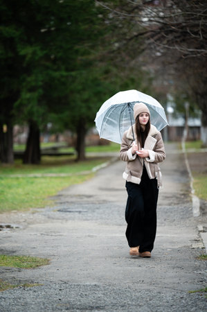 A fashionable young woman strolls down a lush park walkway, holding a transparent umbrella, dressed warmly for a cool and breezy afternoon in nature.の写真素材