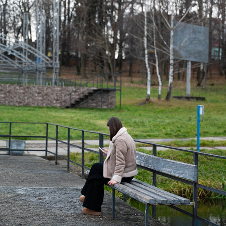 A solitary young woman is focused on her smartphone, sitting on a bench in an empty park. The area features lush greenery with a hint of early spring, creating a serene atmosphere.の写真素材