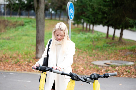 On a cool autumn day, a young woman in a chic white coat checks out electric scooters in a park filled with colorful foliage, ideal for exploration.の写真素材