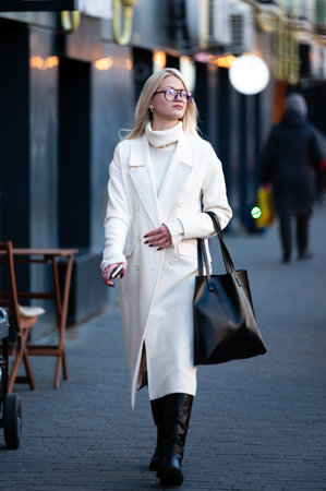A woman in a chic white coat walks confidently along a bustling city street as evening lights twinkle in the background, creating a lively urban atmosphere.の写真素材