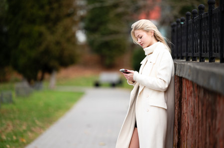 Dressed in a fashionable coat, a young woman quietly engages with her phone while leaning against a textured wall in a peaceful park, with vibrant autumn colors framing the scene.の写真素材