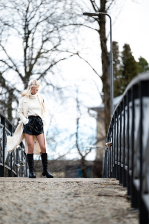 A stylish woman wears a chic outfit as she strolls across a quaint bridge, enjoying the serene surroundings and the freshness of early spring in a vibrant urban area.の写真素材