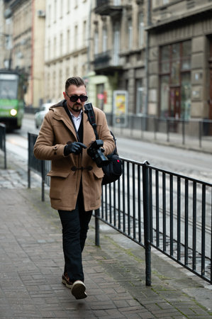 Dressed in a fashionable coat, a man strolls along a bustling city street, camera in hand, ready to capture the essence of urban life in the cold winter air.の写真素材