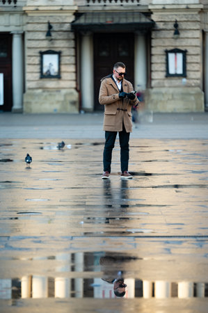 In a tranquil city square, a man stands absorbed in his phone while rainwater creates shimmering reflections, with pigeons wandering peacefully around him as dusk approaches.の写真素材