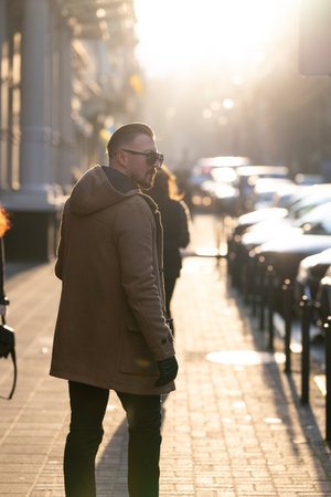 Amidst the golden glow of sunset, a stylish individual in a warm coat walks through a lively street, surrounded by people and parked cars, embodying the seasonal charm.の写真素材