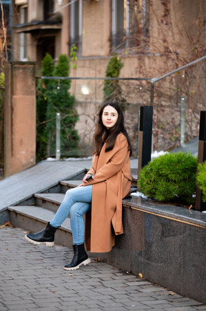 A young woman with long hair sits calmly on stone steps in an urban park. She wears a cozy brown coat and jeans while looking thoughtfully into the distance amid lush greenery.の写真素材