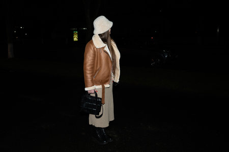 A woman stands confidently in a brown shearling jacket and beige skirt, holding a black handbag as she enjoys a quiet night walk in a park filled with soft city lights.の写真素材