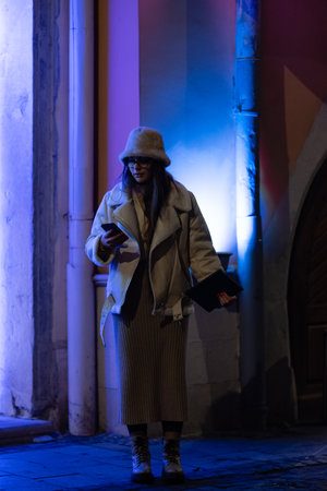 A stylish individual stands on a cobblestone street, illuminated by colorful blue lights, engrossed in their smartphone amidst a lively urban atmosphere.の写真素材
