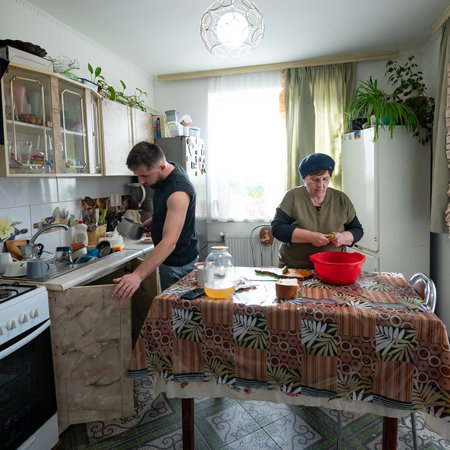 In a warm kitchen filled with sunlight, a young man washes dishes while an elderly woman prepares food, creating a joyful atmosphere of companionship and shared traditions.の写真素材