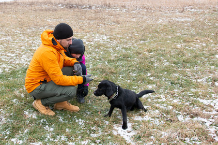 A father crouches down to engage with his playful daughter and their lively black lab puppy in a snowy field, creating joyful memories on a crisp, wintry day.の写真素材