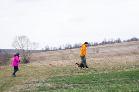 Two individuals stroll through serene countryside fields on a cool day while a dog happily runs alongside them, taking in the beauty of early spring.の写真素材