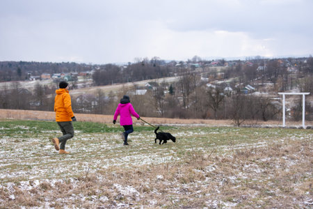 Two individuals walk hand in hand with a black dog on a frosty field, surrounded by a picturesque landscape of countryside homes and rolling hills beneath an overcast sky.の写真素材