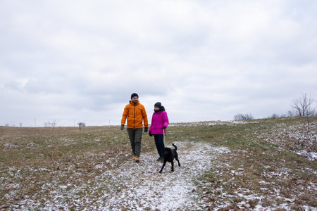Two individuals stroll together on a frosty trail, accompanied by their playful black dog, as winter clouds loom overhead in a serene outdoor setting.の写真素材