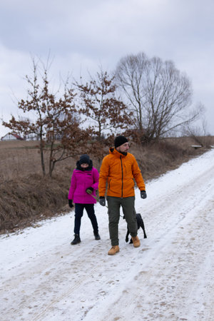 A man and a girl dressed warmly stroll along a snow covered road with a black dog, enjoying the serene winter landscape surrounded by nature.の写真素材