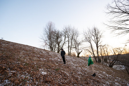 Two individuals traverse a gently sloping hillside covered with fallen leaves and light snow, surrounded by bare trees as the sun sets in a picturesque winter twilight.の写真素材