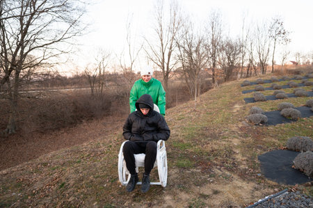 One friend pushes another down a gentle slope in a wheelbarrow, both dressed warmly for the cool weather, surrounded by bare trees and rows of lavender.の写真素材
