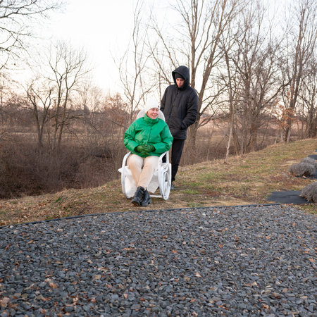 Two friends enjoying a winter adventure by the riverside. One is in a wheelchair while the other pushes with care, surrounded by bare trees and soft sunlight.の写真素材