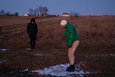 Two individuals enjoy an evening walk in a serene countryside, one observing while the other interacts with a patch of frost on the ground as twilight descends.の写真素材