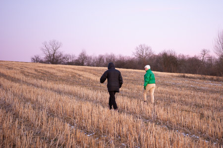 Two people wander a tranquil winter field as dusk settles, appreciating the serene beauty of nature with bare trees and a cool, crisp atmosphere.の写真素材