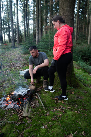 Two friends gather around a warm campfire in a lush forest, preparing to roast food while surrounded by towering trees and a serene atmosphere, creating unforgettable memories.の写真素材
