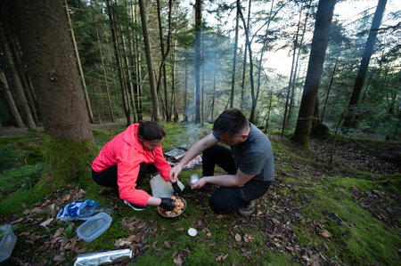 Two friends enjoy cooking over an open fire in a tranquil forest, gathering ingredients and savoring the moment amidst nature's beauty and fresh air.の写真素材