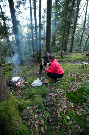 A couple gathers around a small campfire in a lush green forest, surrounded by tall trees as they prepare an outdoor meal, enjoying the peacefulness of nature.の写真素材