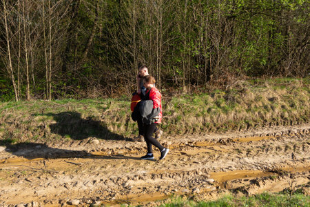 Two individuals stroll along a muddy trail surrounded by vibrant greenery under a bright sun, relishing the beauty of nature during a warm spring afternoon.の写真素材