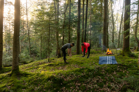 Two friends enjoy a peaceful moment in a vibrant forest, preparing their campsite amid lush greenery and the soothing sounds of nature as the sun begins to set.の写真素材