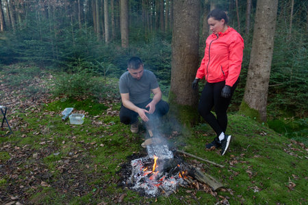 In a tranquil woodland setting, a couple prepares a delicious barbecue over an open flame, surrounded by towering trees and the soothing sounds of nature.の写真素材