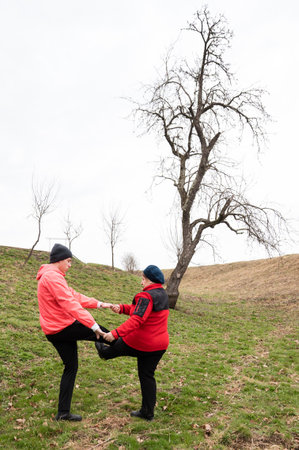 Two friends engage in a playful activity, lifting their legs in a grassy field while sharing smiles, surrounded by the tranquility of early spring with a cloudy sky overhead.の写真素材
