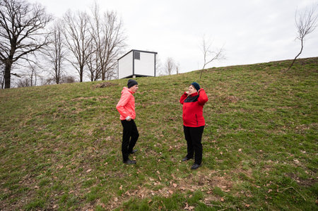 Two people chat on a grassy incline, dressed warmly for the chilly weather, while a minimalist building stands nearby under an overcast sky.の写真素材