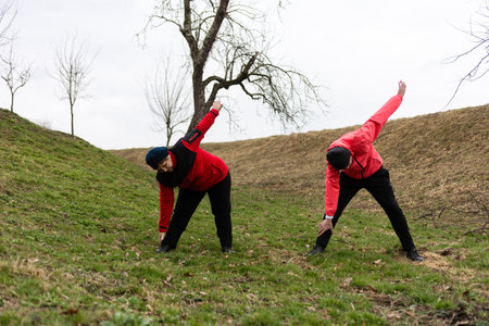 Two individuals in red jackets stretch on a grassy slope under a cloudy sky, embracing a healthy morning routine surrounded by nature's tranquility during early spring.の写真素材