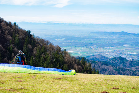 A paraglider readies equipment on a grassy mountain slope, surrounded by vibrant greenery and a panoramic view of valleys stretching to the horizon under clear skies.の写真素材