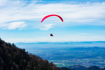 High above a lush valley, a paraglider with a vibrant red canopy glides gracefully through the sky, surrounded by majestic mountains and breathtaking views on a clear day.の写真素材