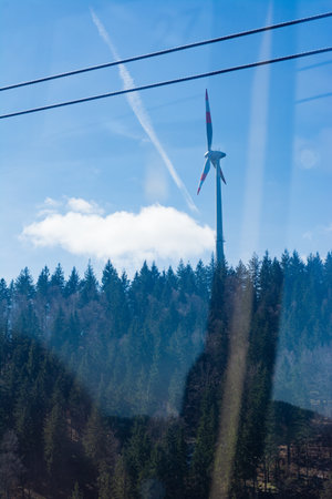 A vibrant wind turbine rises amidst tall, dense trees under a clear blue sky, with wispy clouds adding charm to the peaceful scenery on a sunny day.の写真素材